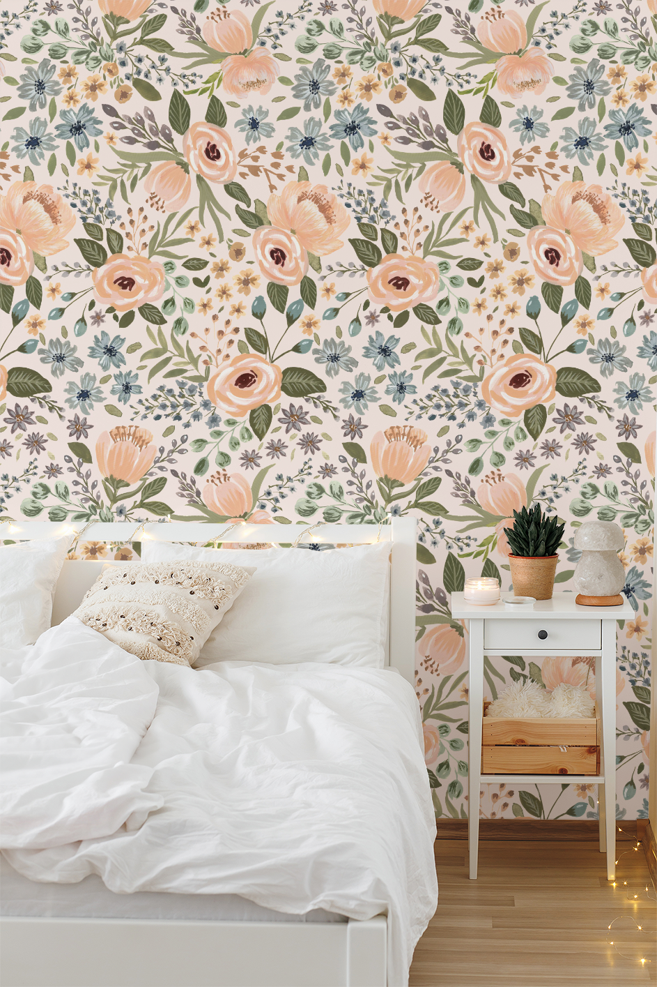 Cozy bedroom corner with white bedding and a nightstand set against Juliette’s Summer Blooms wallpaper, featuring layered watercolor florals in peach, soft blue, and sage green.