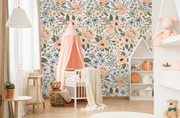 Nursery with oversized peach and blue floral wallpaper behind a white canopy crib, styled with warm wood accents and soft textiles, featuring Juliette’s Summer Blooms watercolor wallpaper.