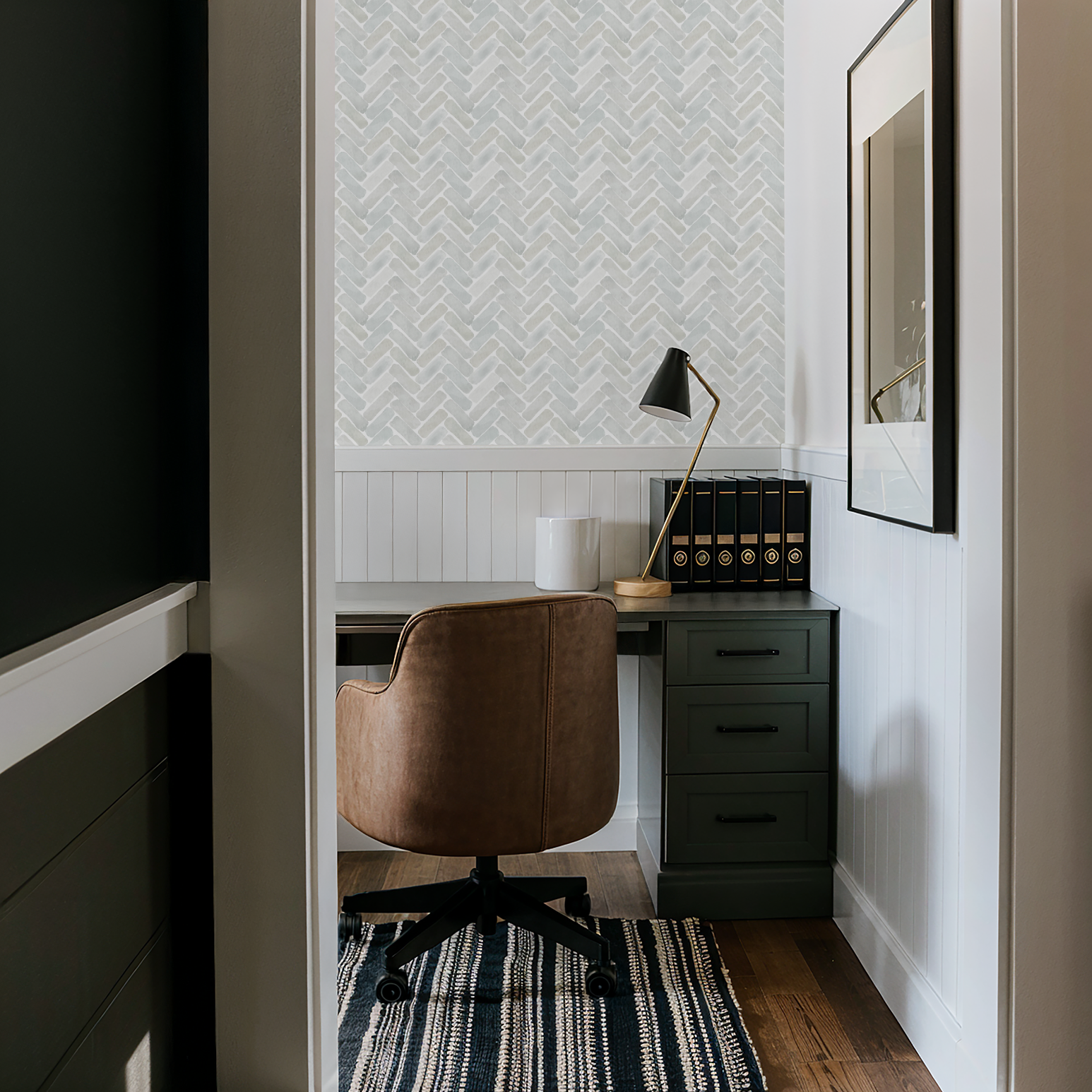 Neutral watercolor herringbone wallpaper in a cozy office nook with beadboard paneling, a leather desk chair, and green cabinetry.