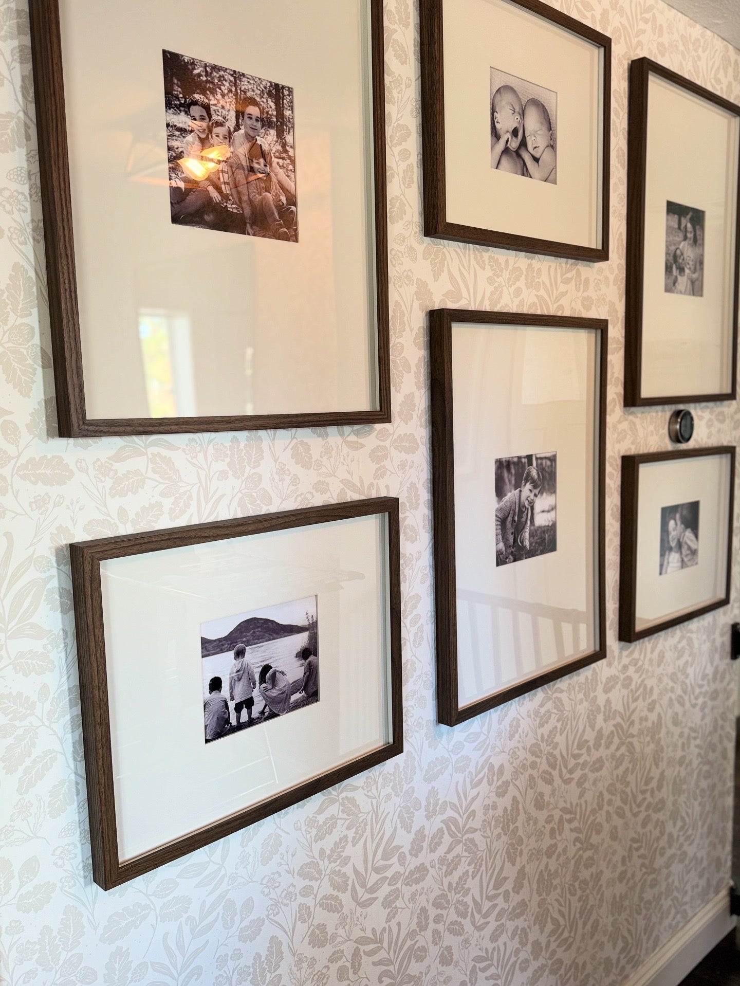 Pale oak botanical wallpaper behind a gallery wall of wood-framed family photos in a hallway with natural light.