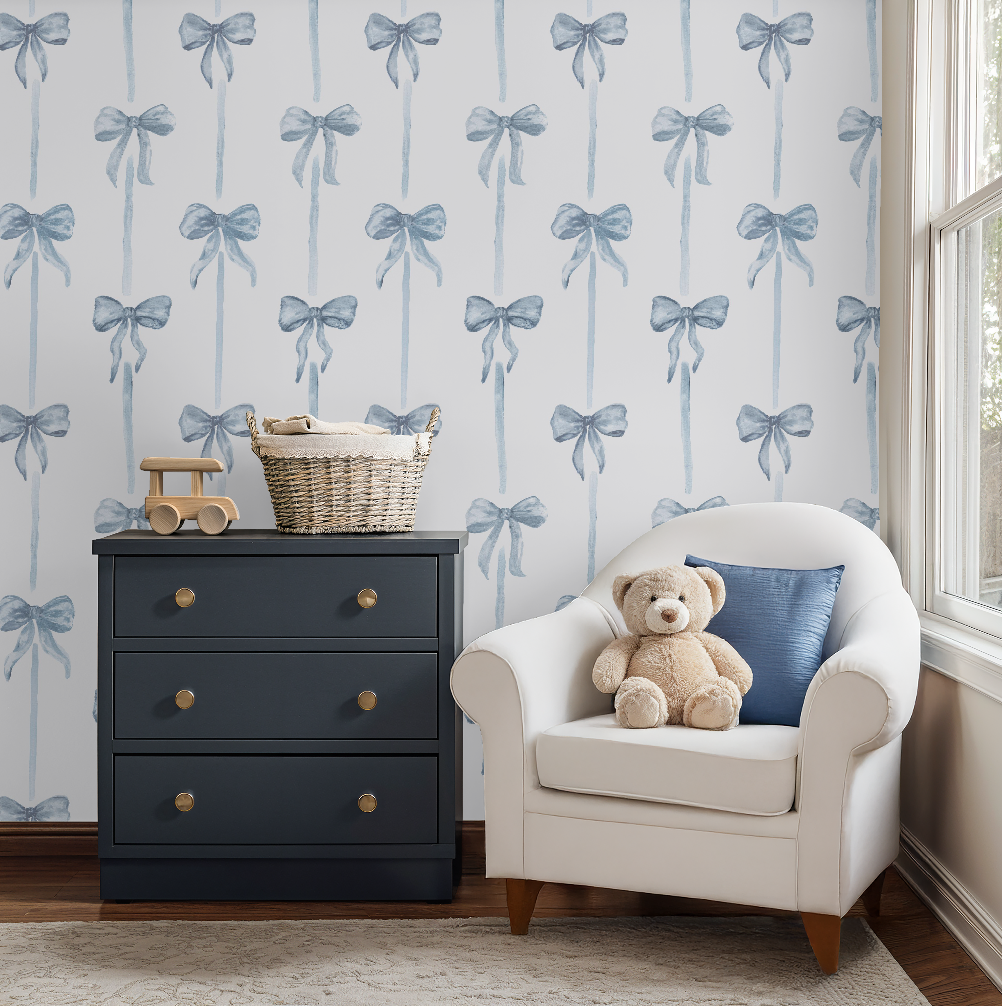 Nursery room with blue bow-patterned wallpaper, dark gray dresser, and white chair with teddy bear and pillow.