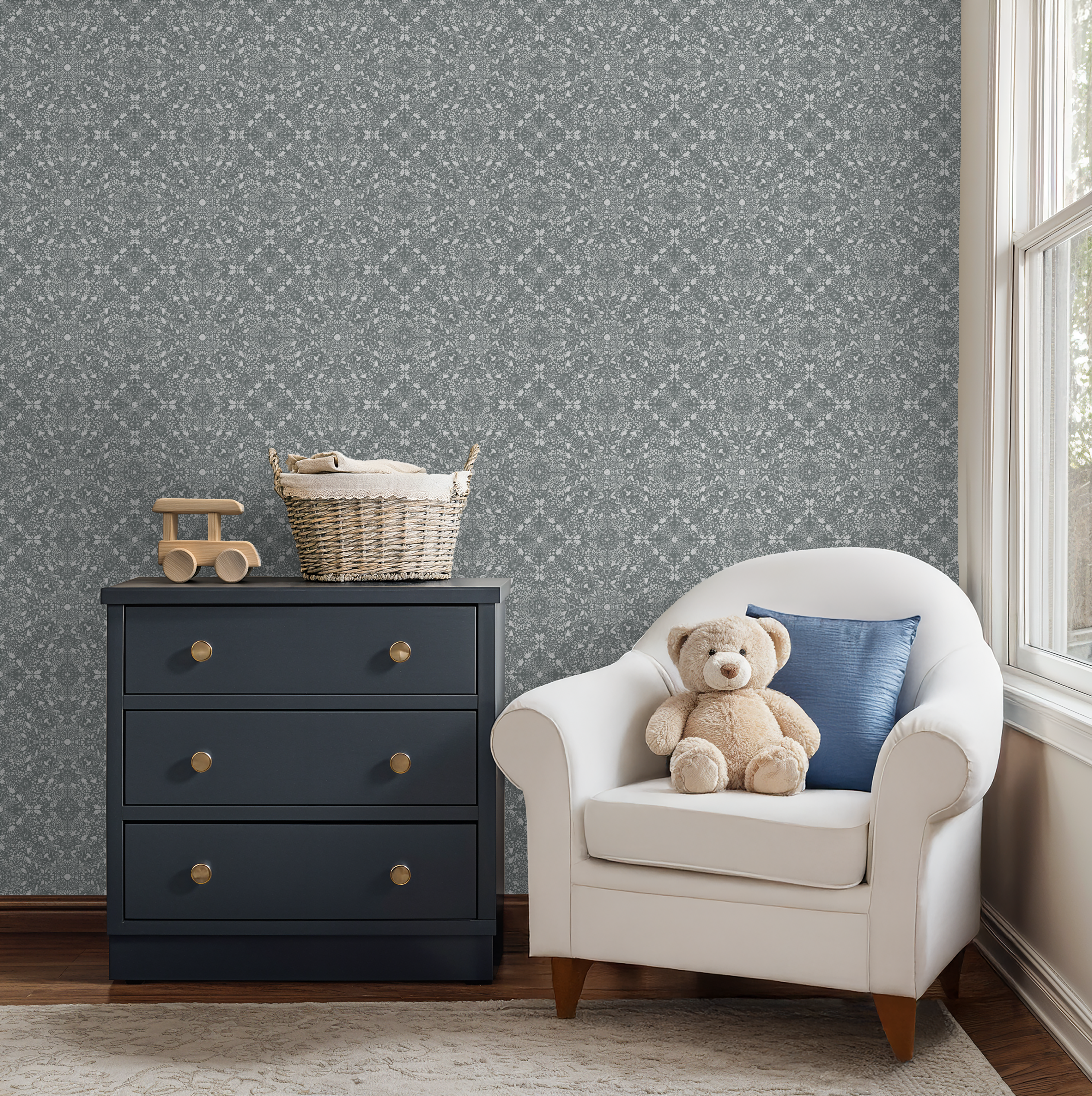 Nursery corner featuring a navy dresser, plush teddy bear on a white armchair, and Woodland Lace in Blue wallpaper behind.