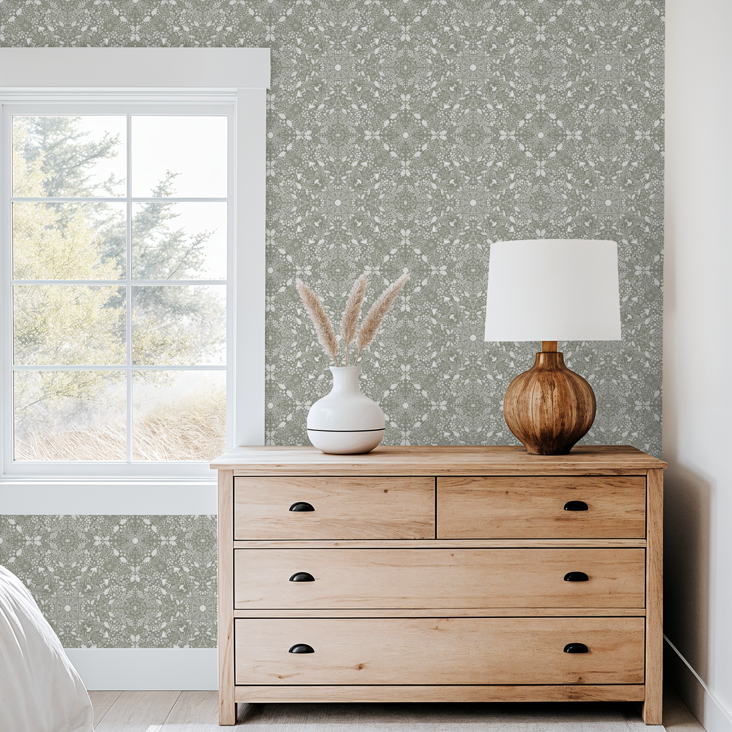 Wooden dresser with a lamp and vase against a patterned Woodland Lace in Sage wallpaper.
