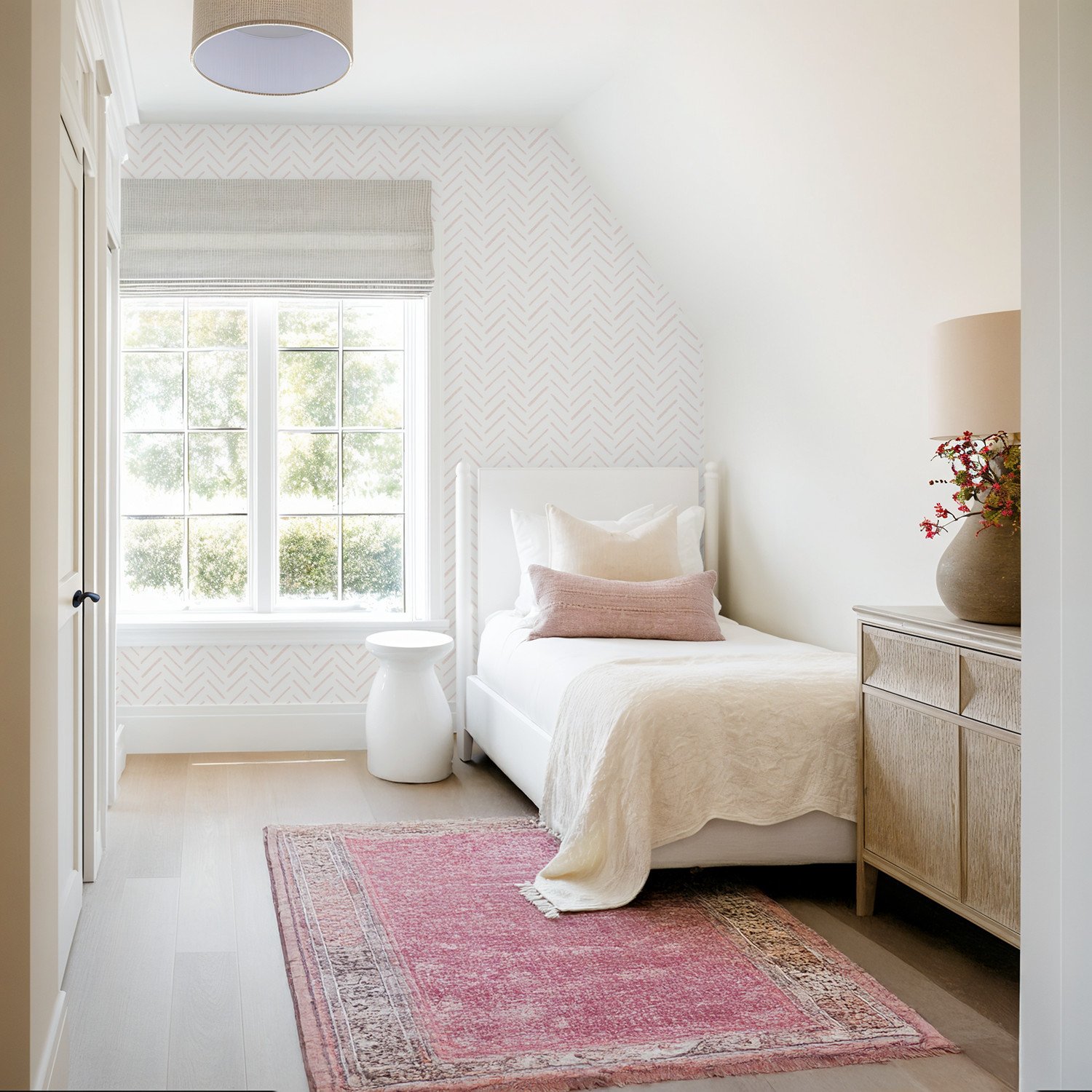 Blush pink herringbone wallpaper in a bright bedroom with a white bed, blush accents, and a pink vintage-style rug.