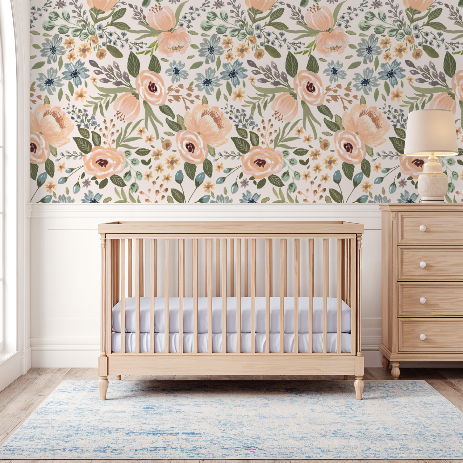 Soft, airy nursery with a natural wood crib and blue rug against Juliette’s Summer Blooms wallpaper, featuring hand-painted florals in peach, golden yellow, and soft blue.