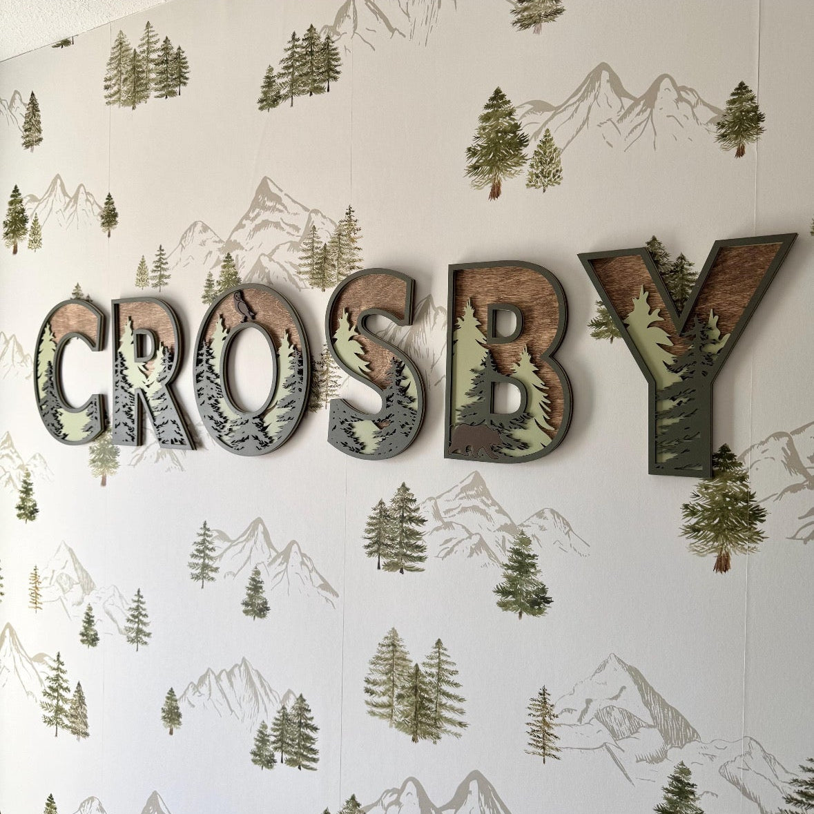 Close-up of ‘Crosby’ wooden name letters mounted on watercolor mountain and evergreen wallpaper.