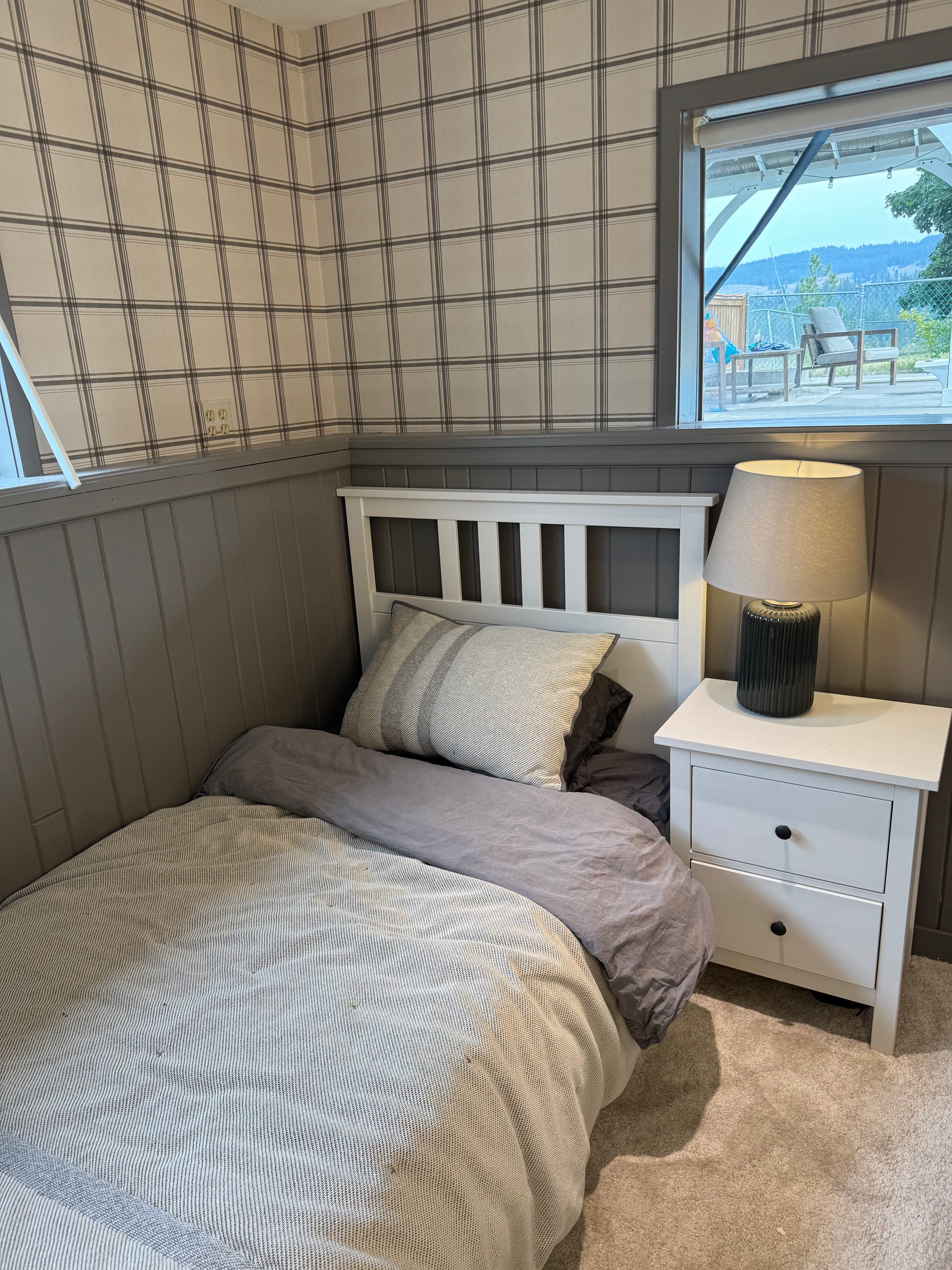 A single bed with beige and gray bedding in a bedroom with gray wood-paneled walls, a white nightstand with a lamp, and a window showing an outdoor view.
