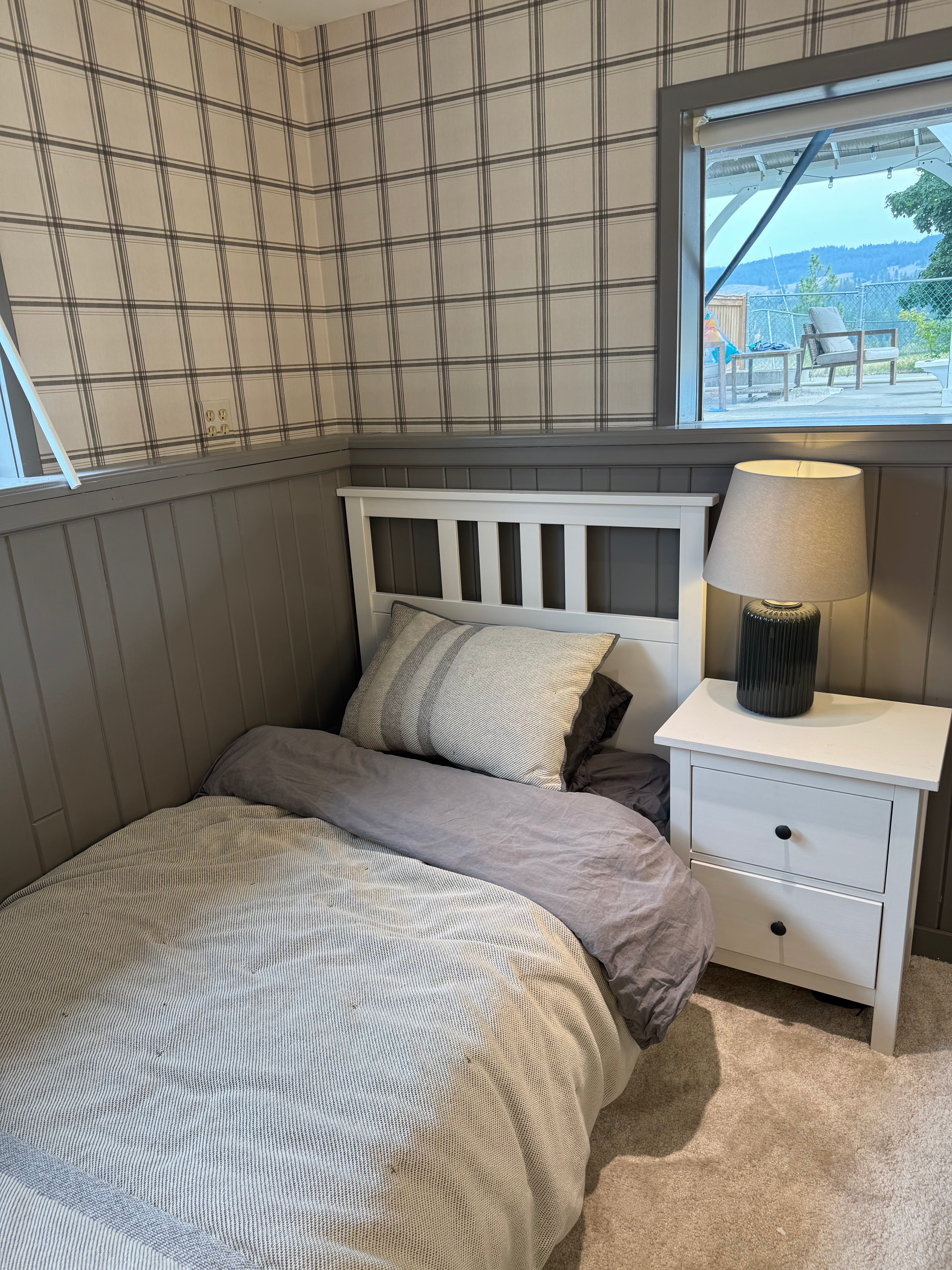 A single bed with beige and gray bedding in a bedroom with gray wood-paneled walls, a white nightstand with a lamp, and a window showing an outdoor view.
