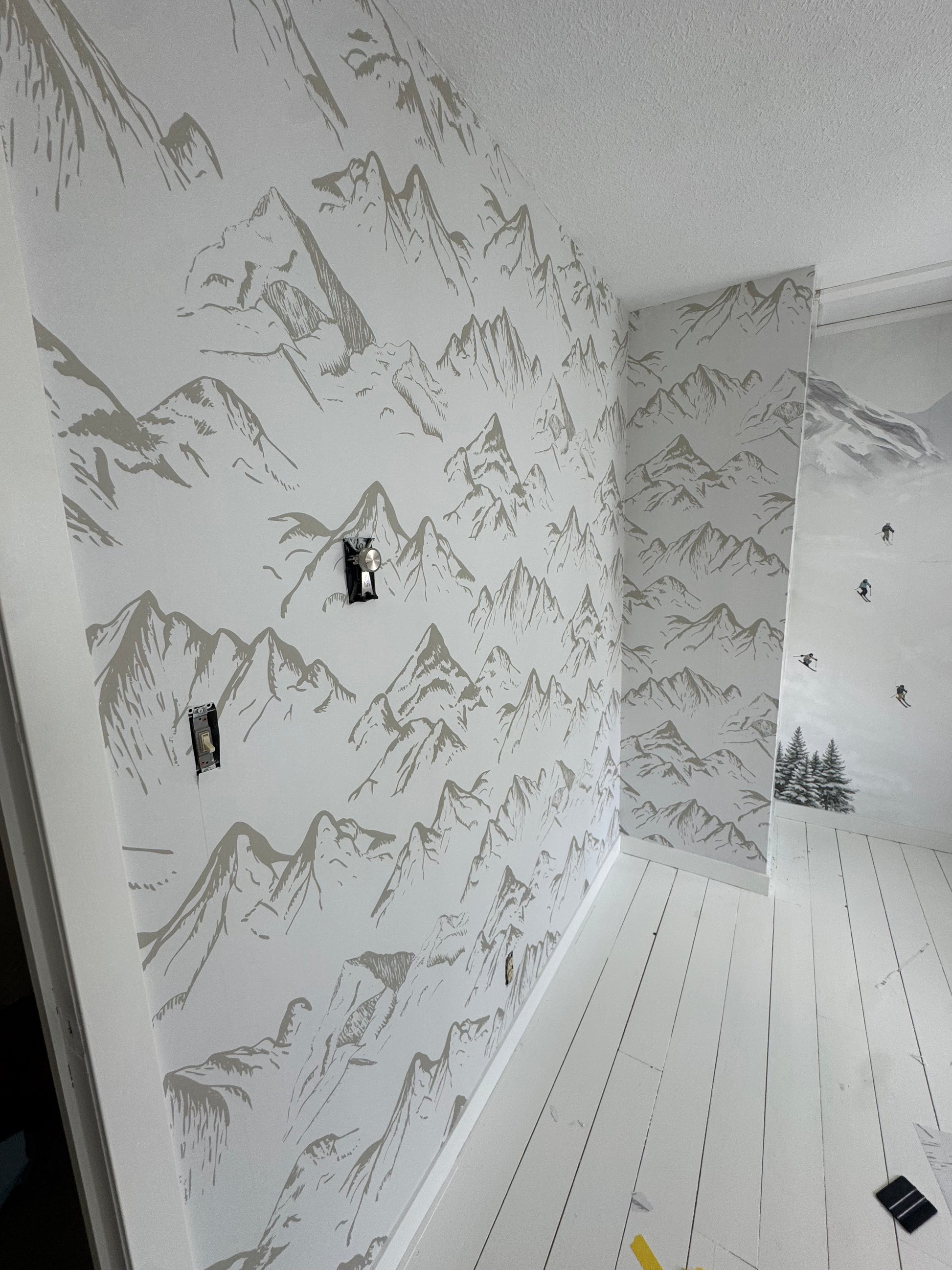 Neutral taupe mountain wallpaper on an angled wall during installation, with outlets exposed and white wood flooring.