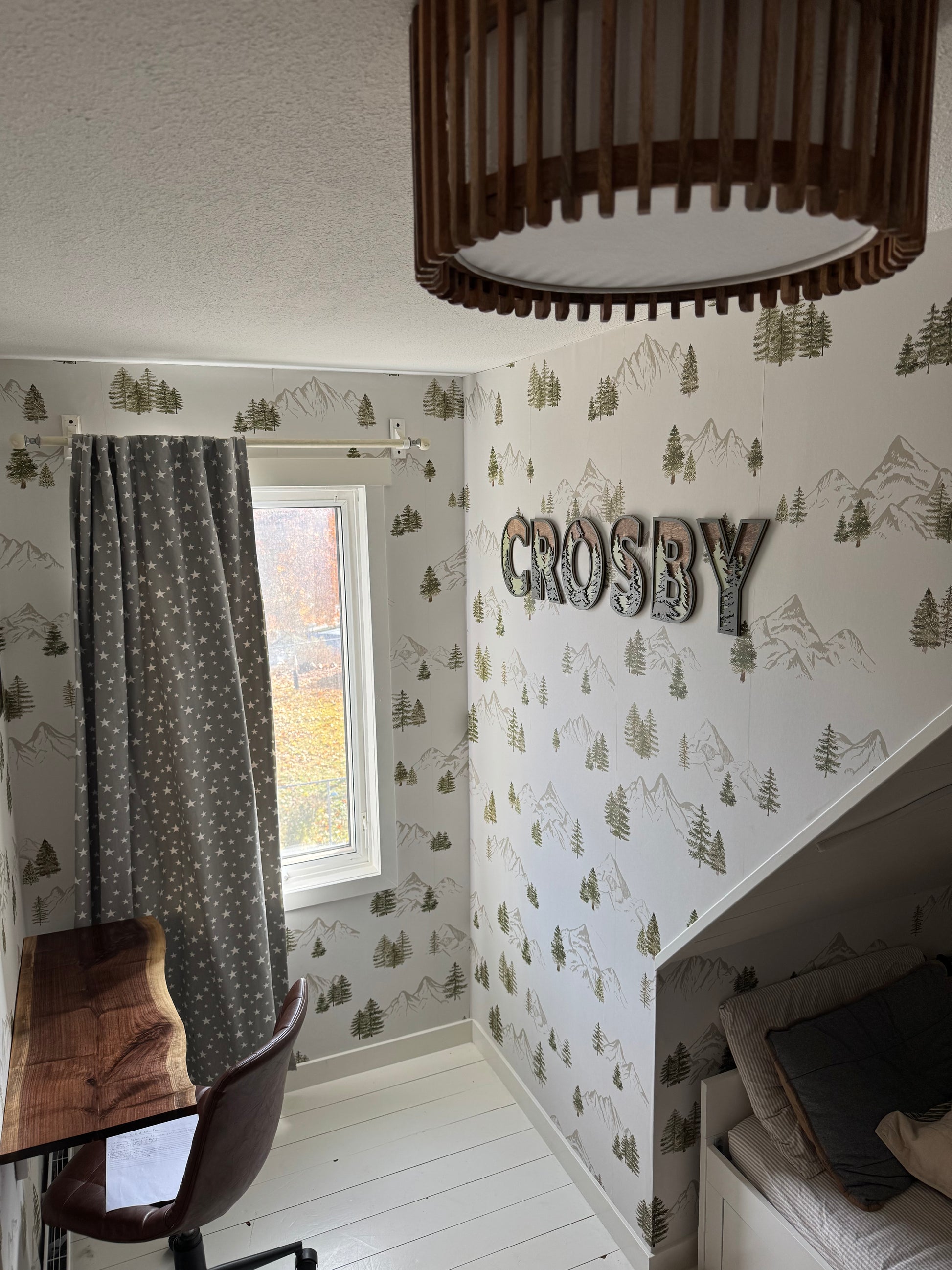 Watercolor mountain and evergreen wallpaper with a custom ‘Crosby’ name sign beside a window with star-patterned curtains.