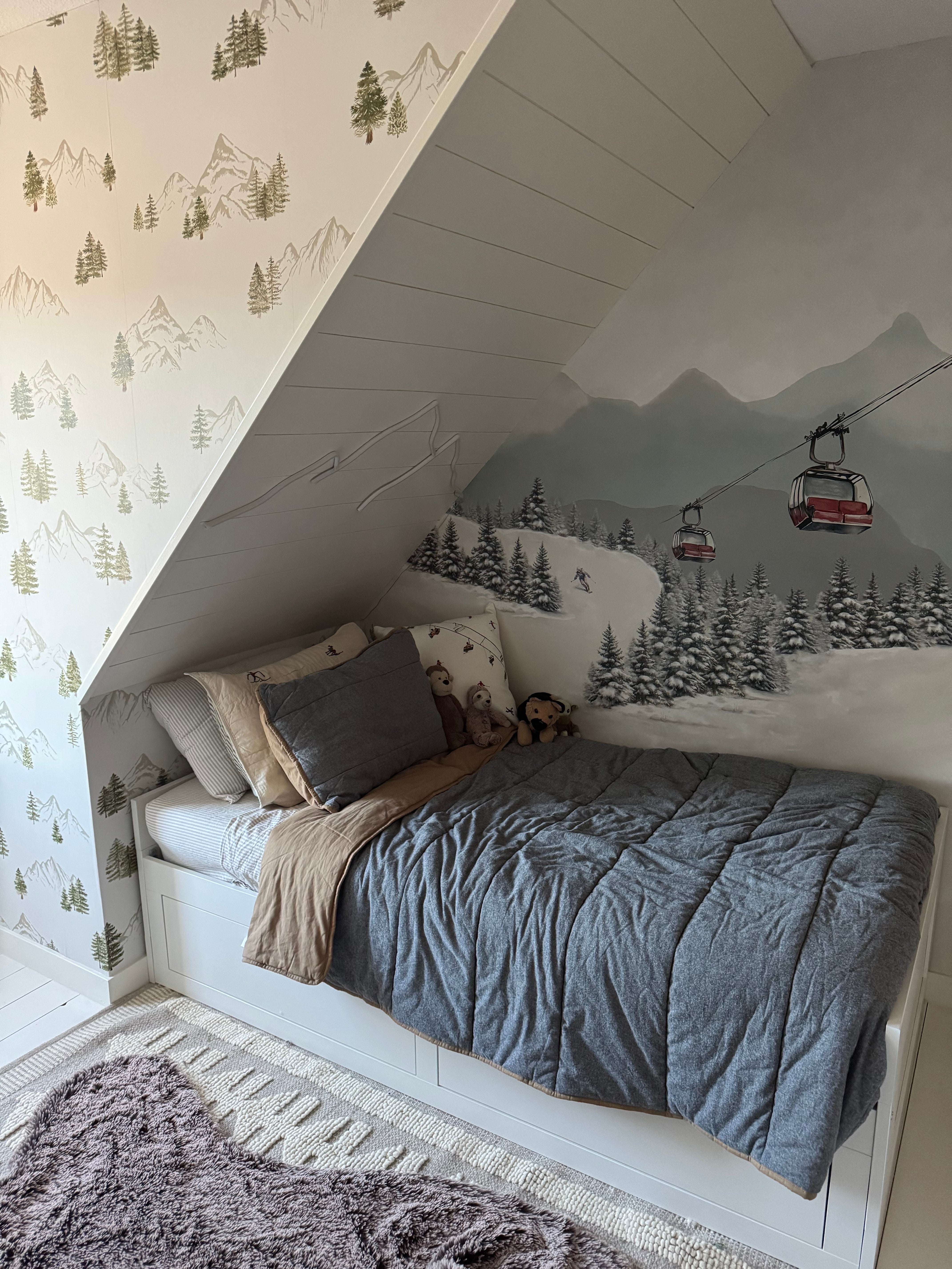 Watercolor alpine ski mountain mural with red gondolas styled under a sloped ceiling beside a coordinating mountain-and-pine wallpaper.