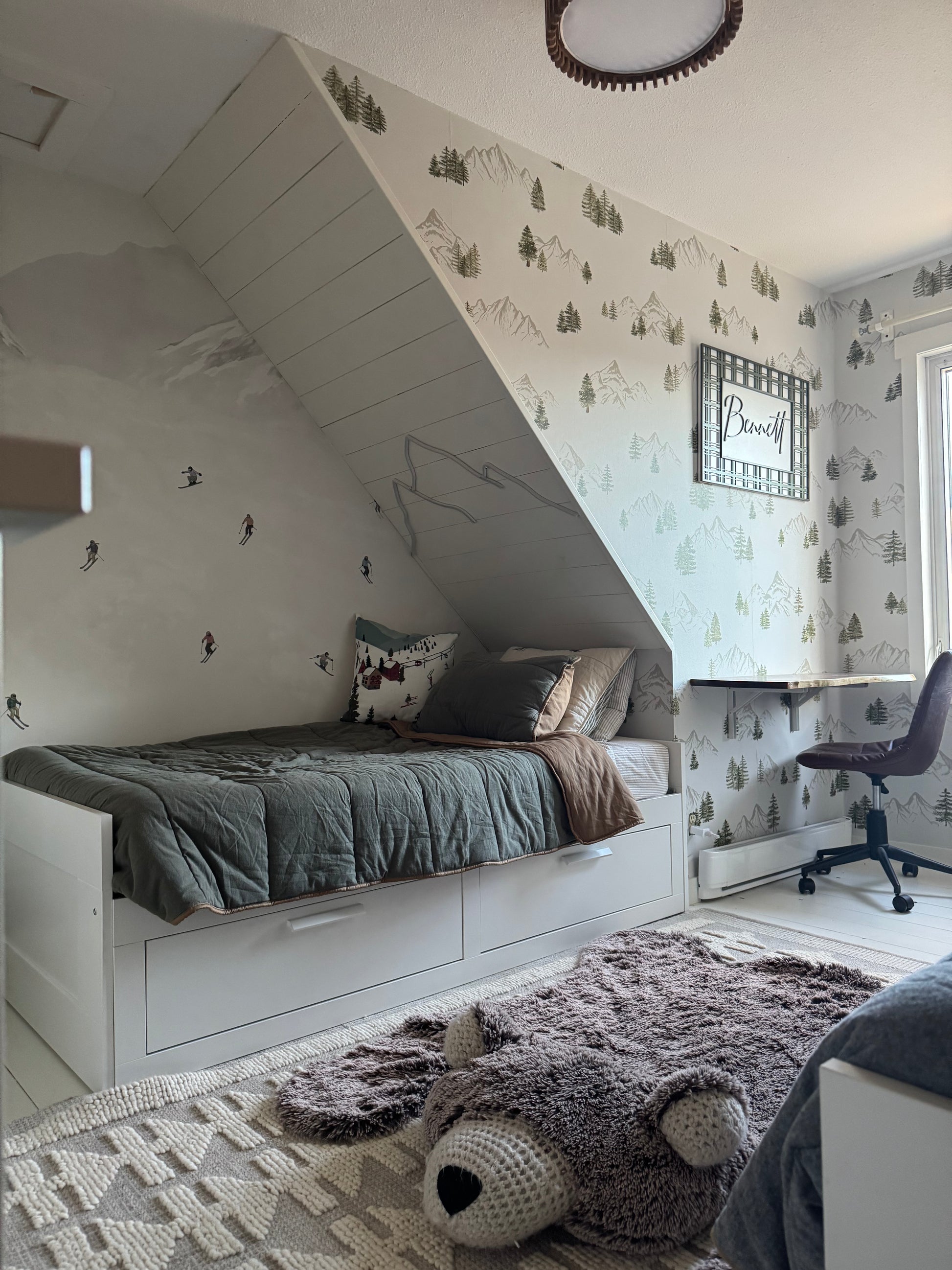 Watercolor mountain and evergreen wallpaper styled under a sloped ceiling beside a cozy bed with neutral bedding.