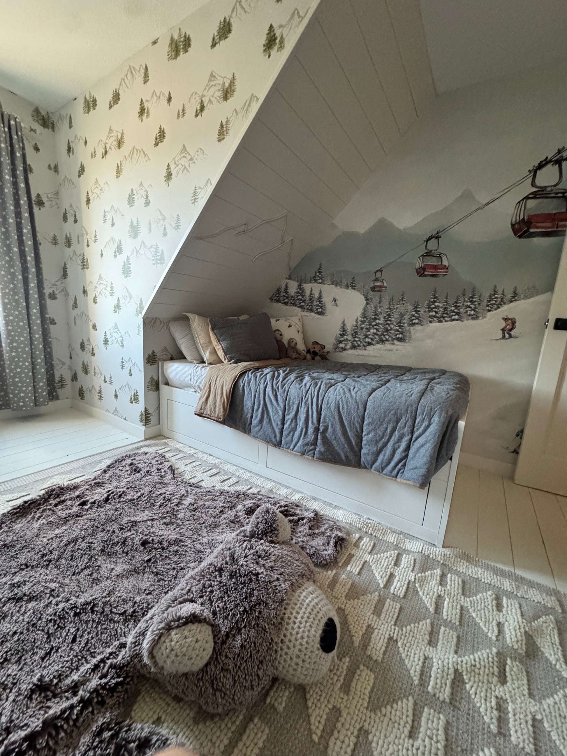 Alpine ski mural with skiers and red gondolas behind a cozy bed and soft bear-shaped rug in a children’s room.