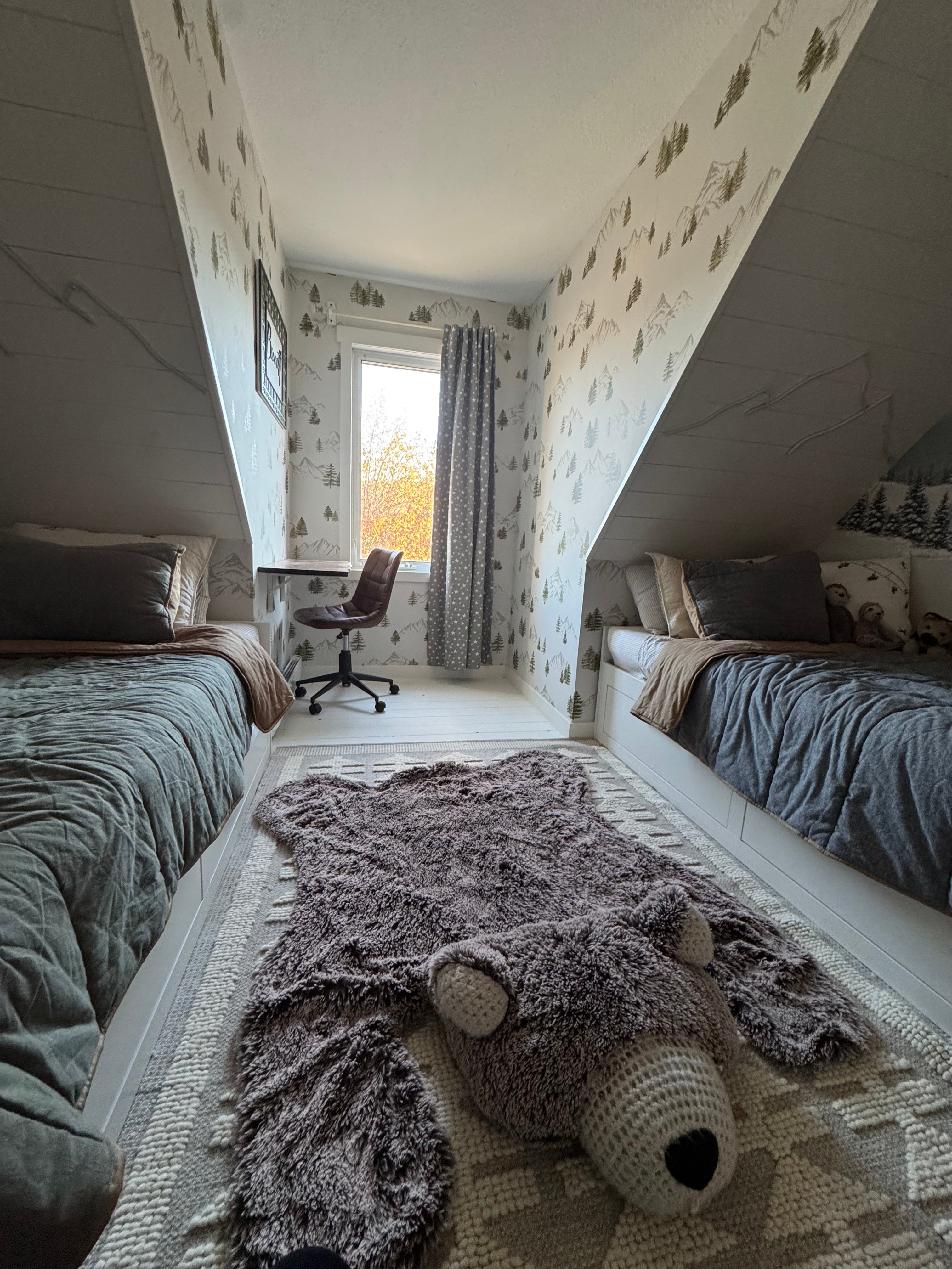 Kids’ room with two beds under angled ceilings, featuring watercolor mountain and evergreen wallpaper and a plush bear-shaped rug.