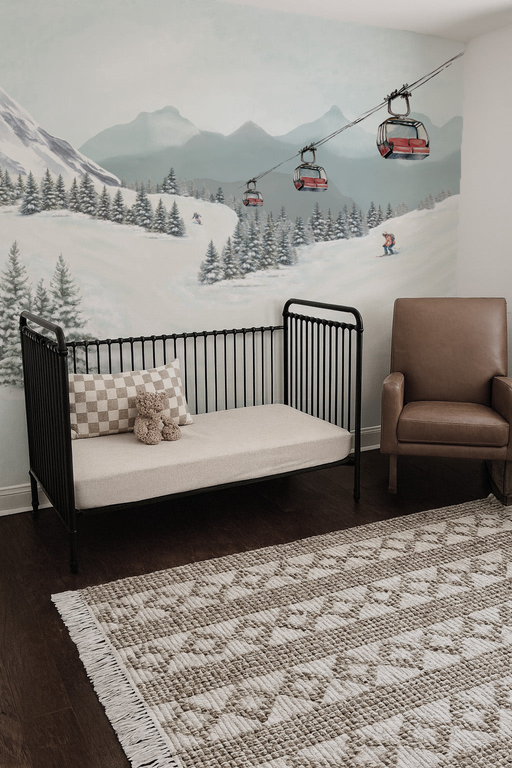 Nursery room with a crib, chair, and mountain mural on the wall depicting a ski resort and chair lift.