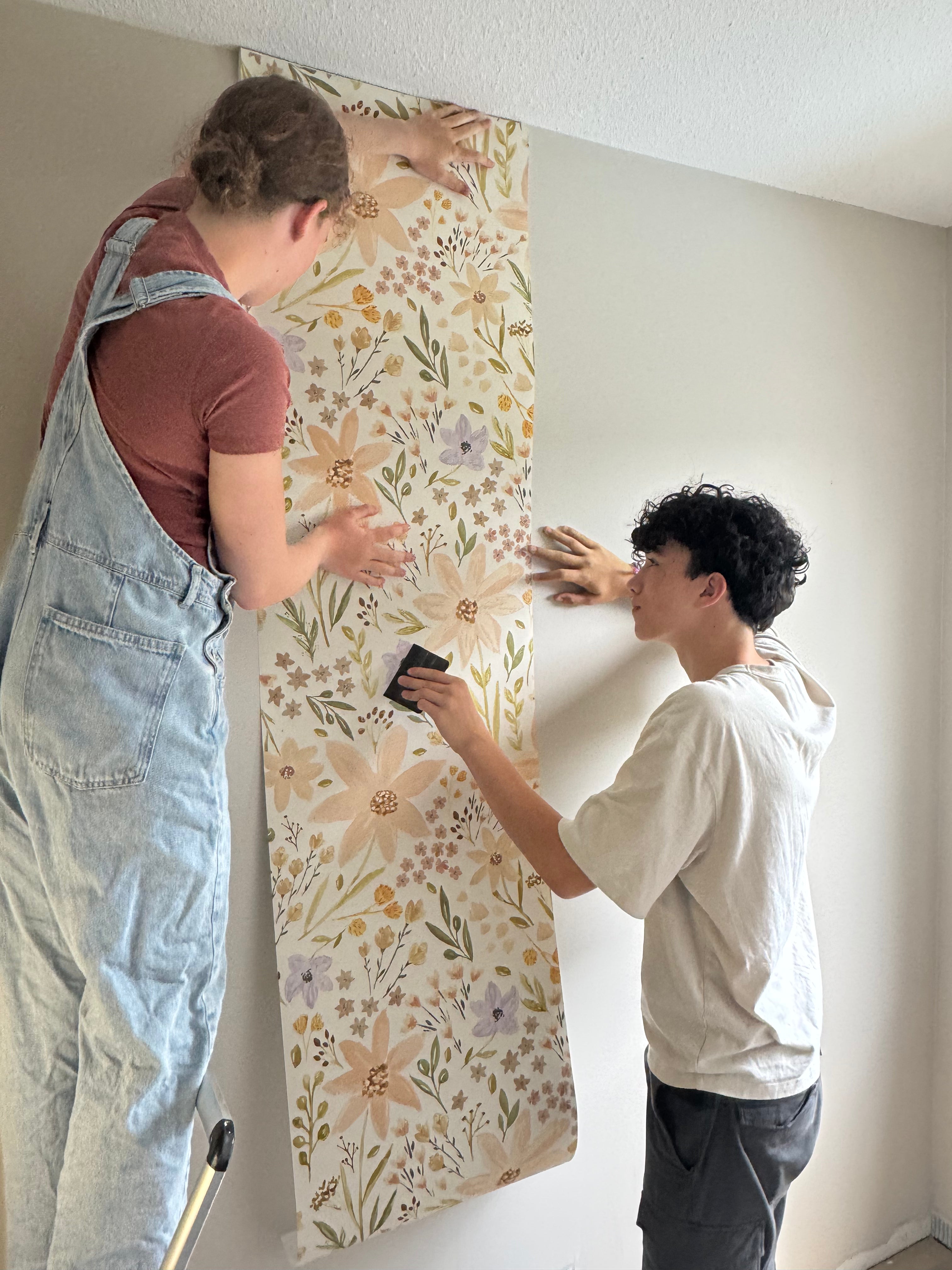 Two people working together to apply floral wallpaper to a wall.