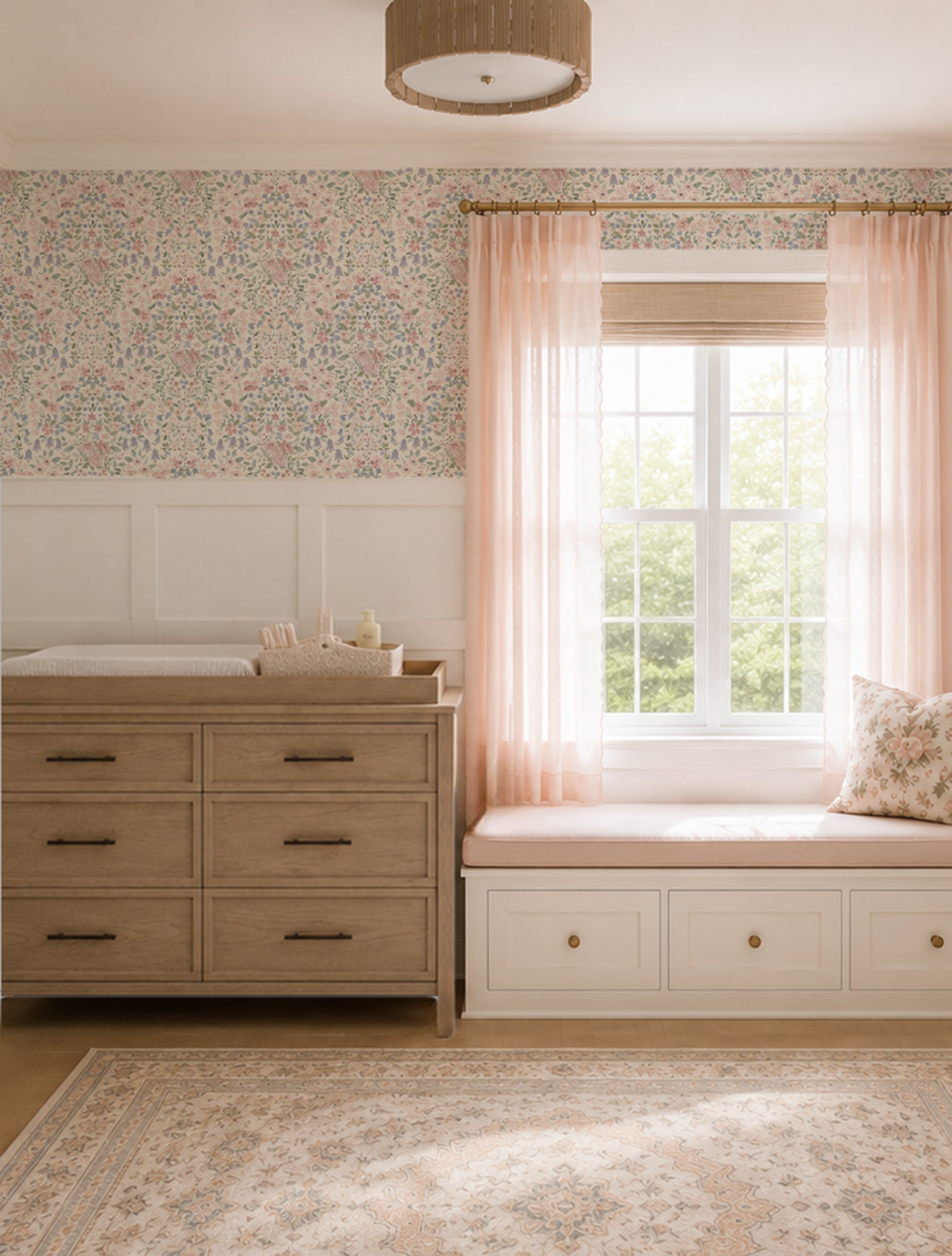 A styled nursery or baby room interior featuring a light wood dresser with a changing pad, a built-in window seat with blush pink curtains, floral wallpaper, and a patterned area rug.
