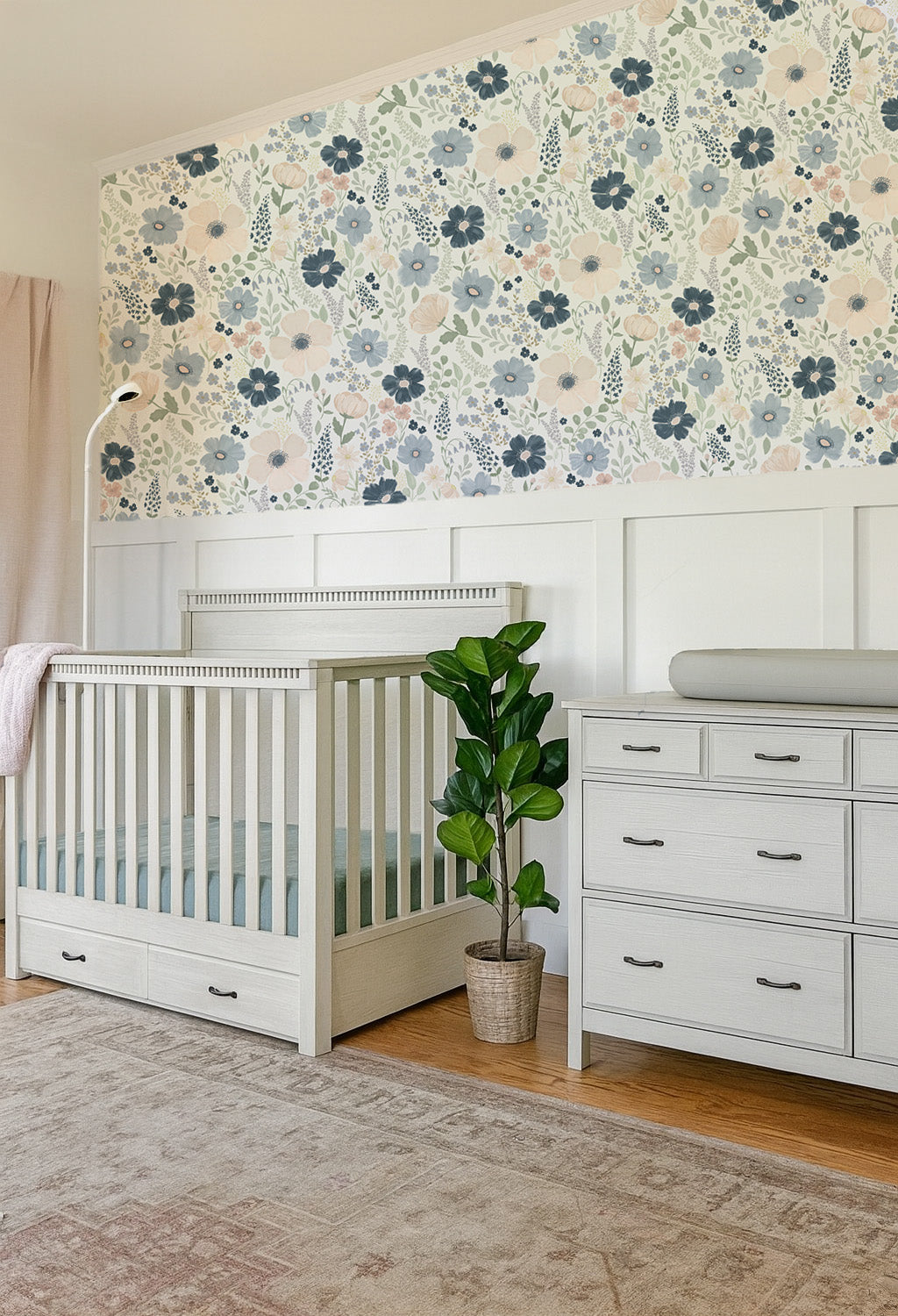 Navy and blush floral wallpaper styled above white paneling in a light nursery with a gray crib, changing dresser, and potted plant.