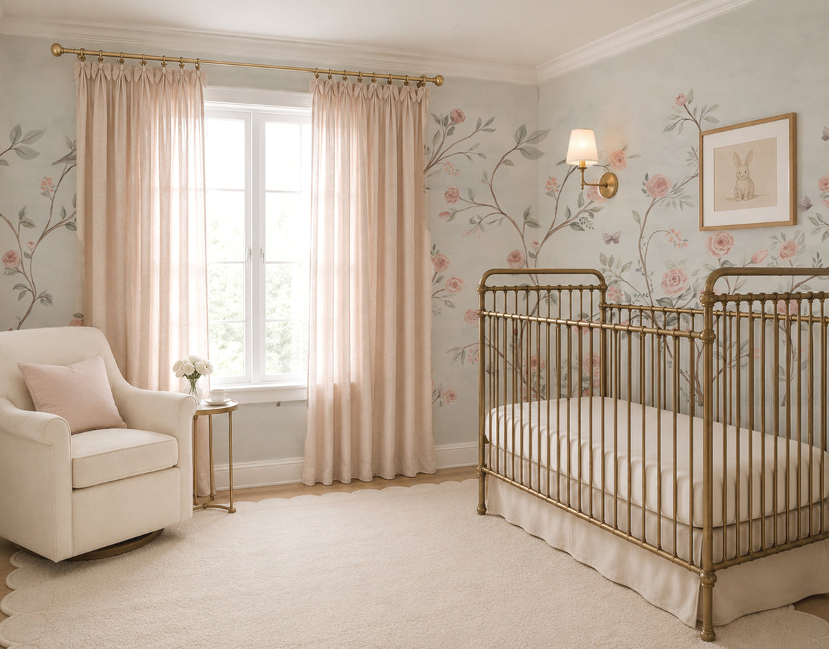 A nursery room featuring a gold metal crib, blush pink floral chinoiserie wallpaper with watercolor roses, and matching blush pink curtains hanging from a gold rod. A cream armchair and a small side table are positioned next to the window.