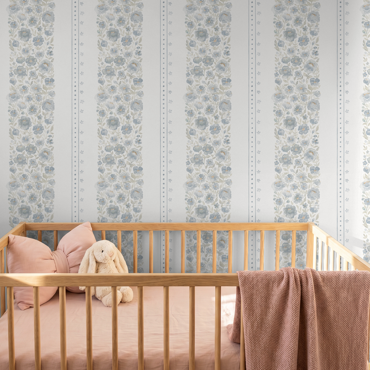 Chambray Cottage blue floral stripe wallpaper behind a natural wood crib styled with soft blush bedding and a plush bunny.