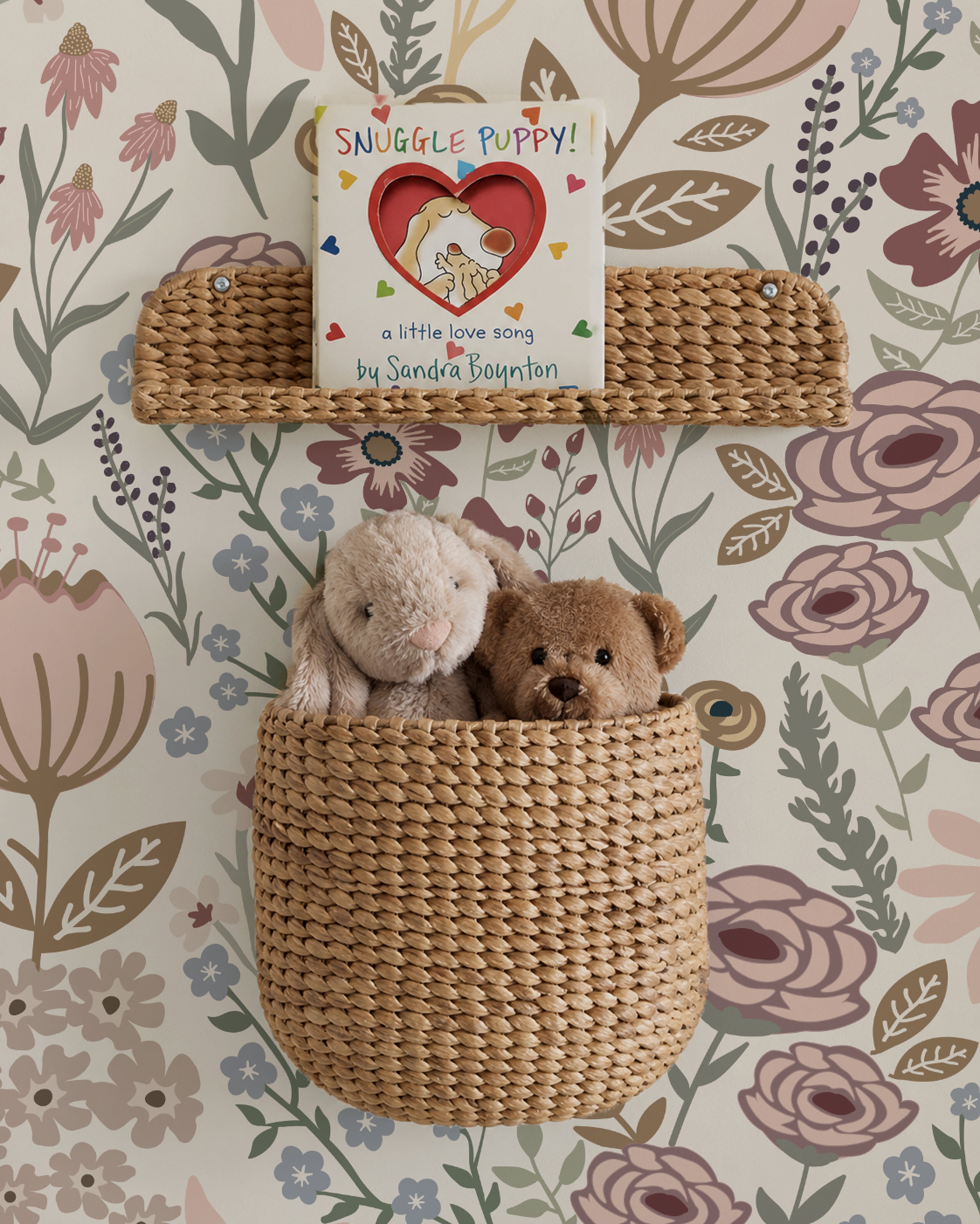 A styled nursery scene featuring two woven wicker baskets mounted on a floral wallpaper background. The top basket holds a children's book titled 'Snuggle Puppy! by Sandra Boynton', and the bottom basket is a larger pocket-style basket containing two plush stuffed animals, a cream bunny and a brown puppy.