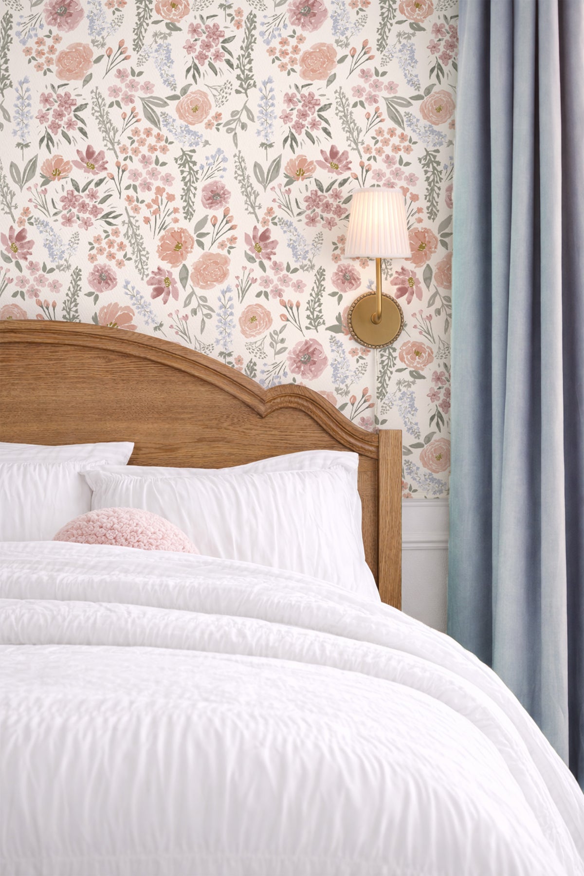 A styled bedroom corner showing a wooden bed headboard with white bedding, floral wallpaper in pink, beige, and green, a wall-mounted lamp, and a blue-gray curtain.