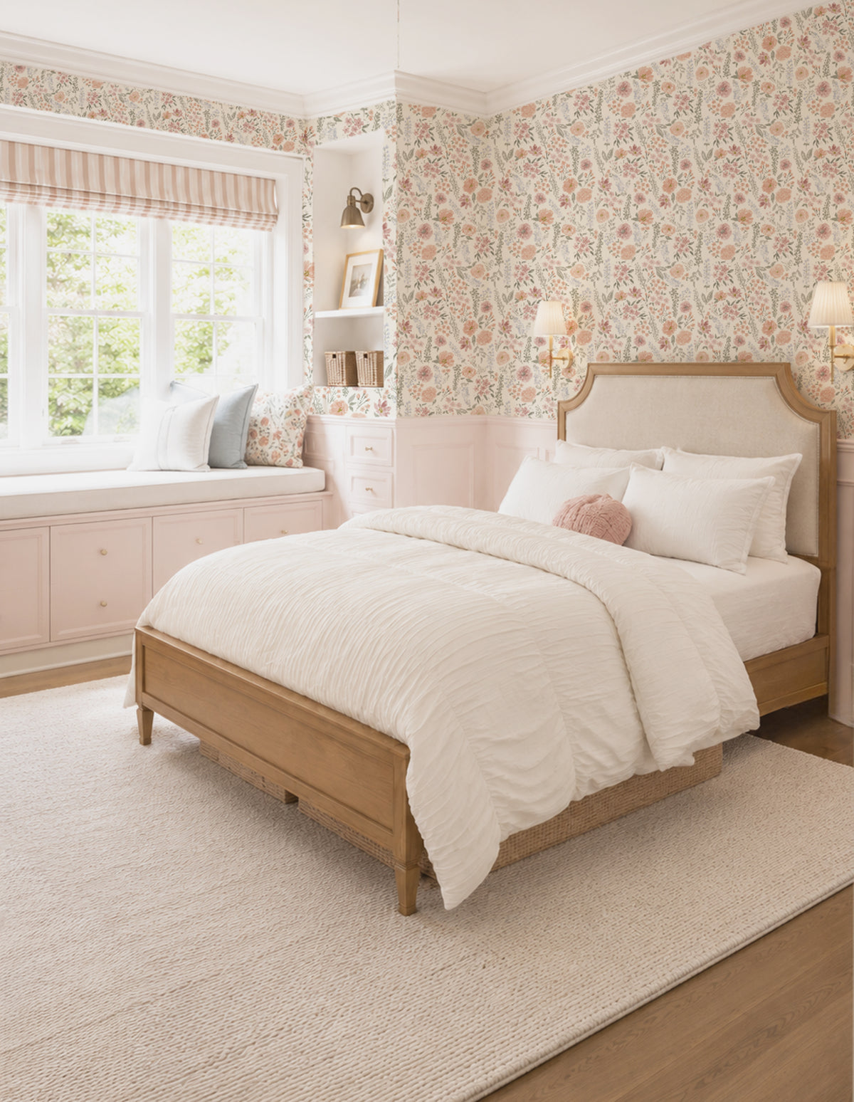 A light wood bed with a cream upholstered headboard, dressed in white bedding and pillows, set in a beautifully decorated bedroom with floral wallpaper, a window seat, and a cream rug.