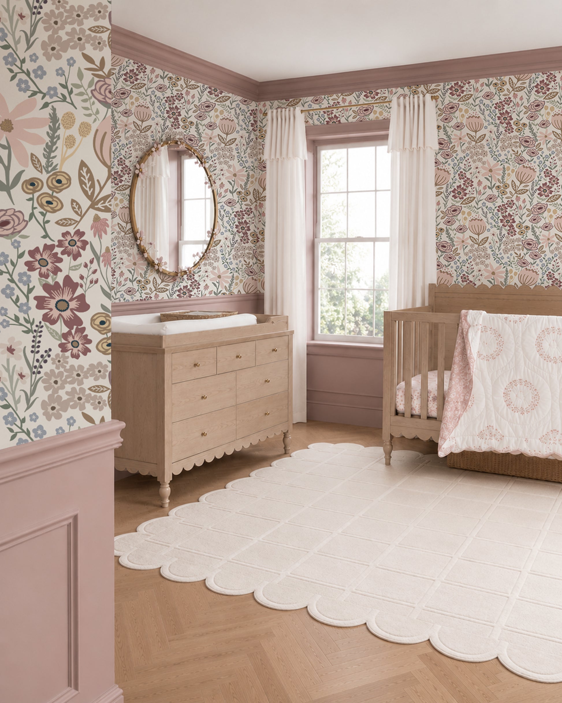 A styled nursery room featuring floral wallpaper with a botanical pattern in muted pink, beige, and green tones. The room includes a light wood dresser with a white changing tray, a round gold-framed mirror, a window with white curtains, a wooden crib with a pink patterned quilt, and a large scalloped cream rug on a light wood herringbone floor.