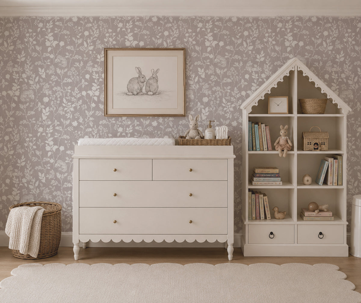 A styled nursery interior featuring a white wooden dresser with scalloped trim, a white house-shaped bookshelf filled with books and toys, patterned wallpaper, a framed bunny print, and a woven basket on the floor.