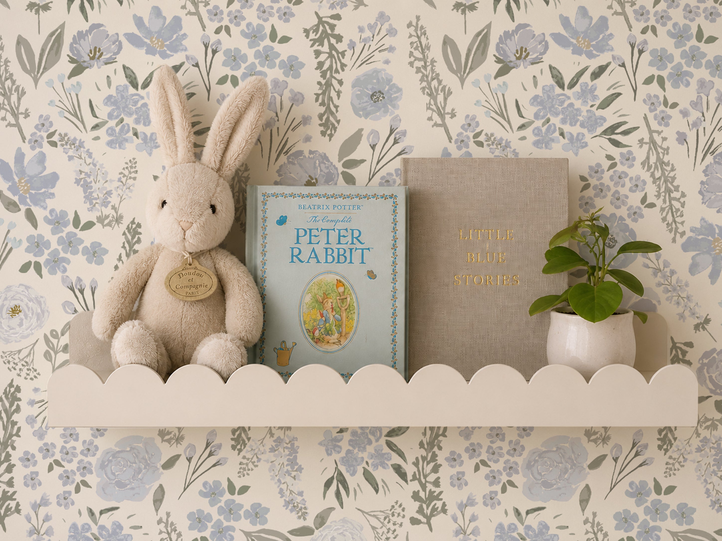 A styled interior scene showing a cream plush rabbit toy, a Peter Rabbit children's book, a beige book with gold lettering, and a small potted green plant arranged on a white scalloped shelf against floral wallpaper.