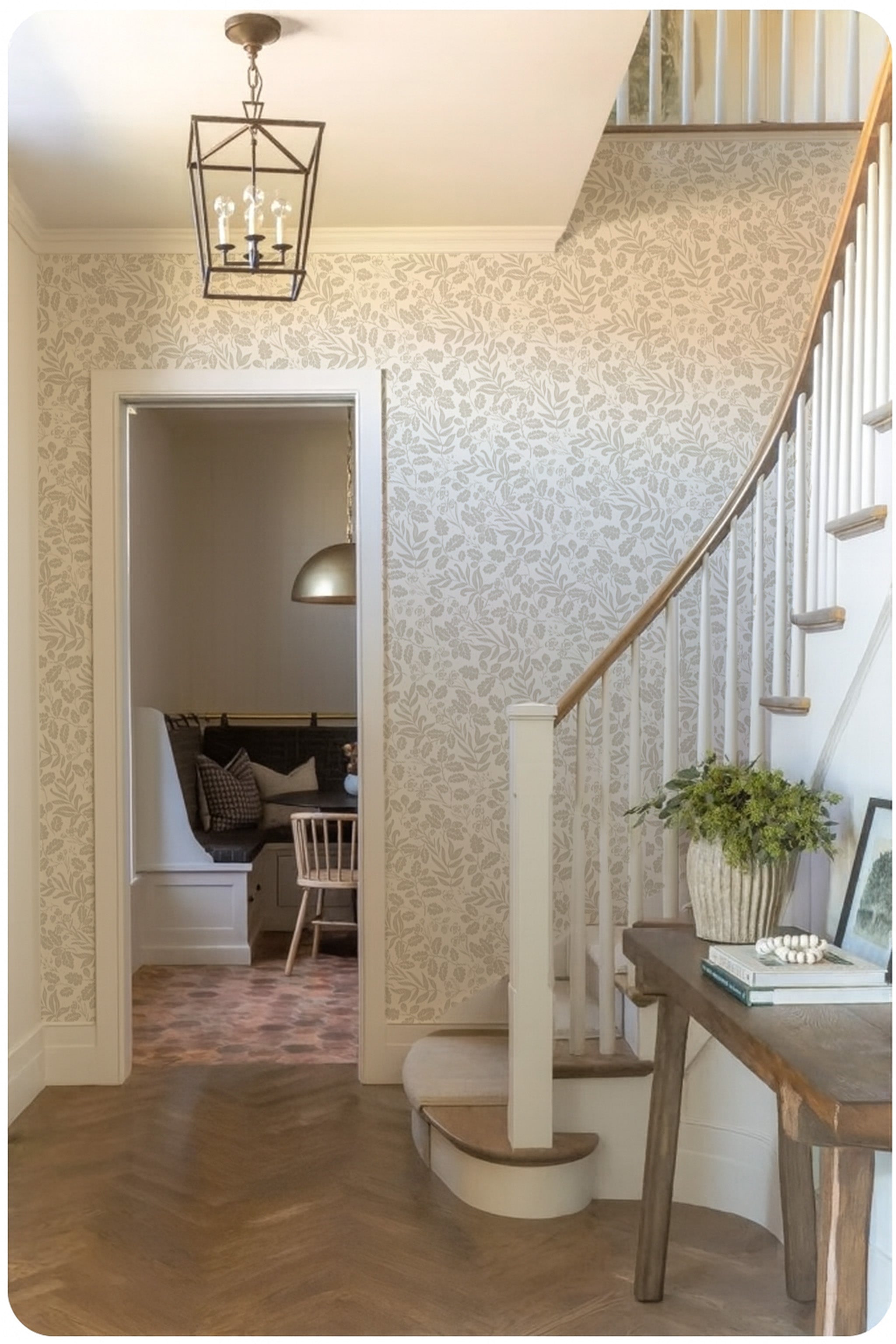 Neutral botanical oak-and-acorn wallpaper in a bright entryway with a curved staircase, wood flooring, and a rustic console table with greenery.