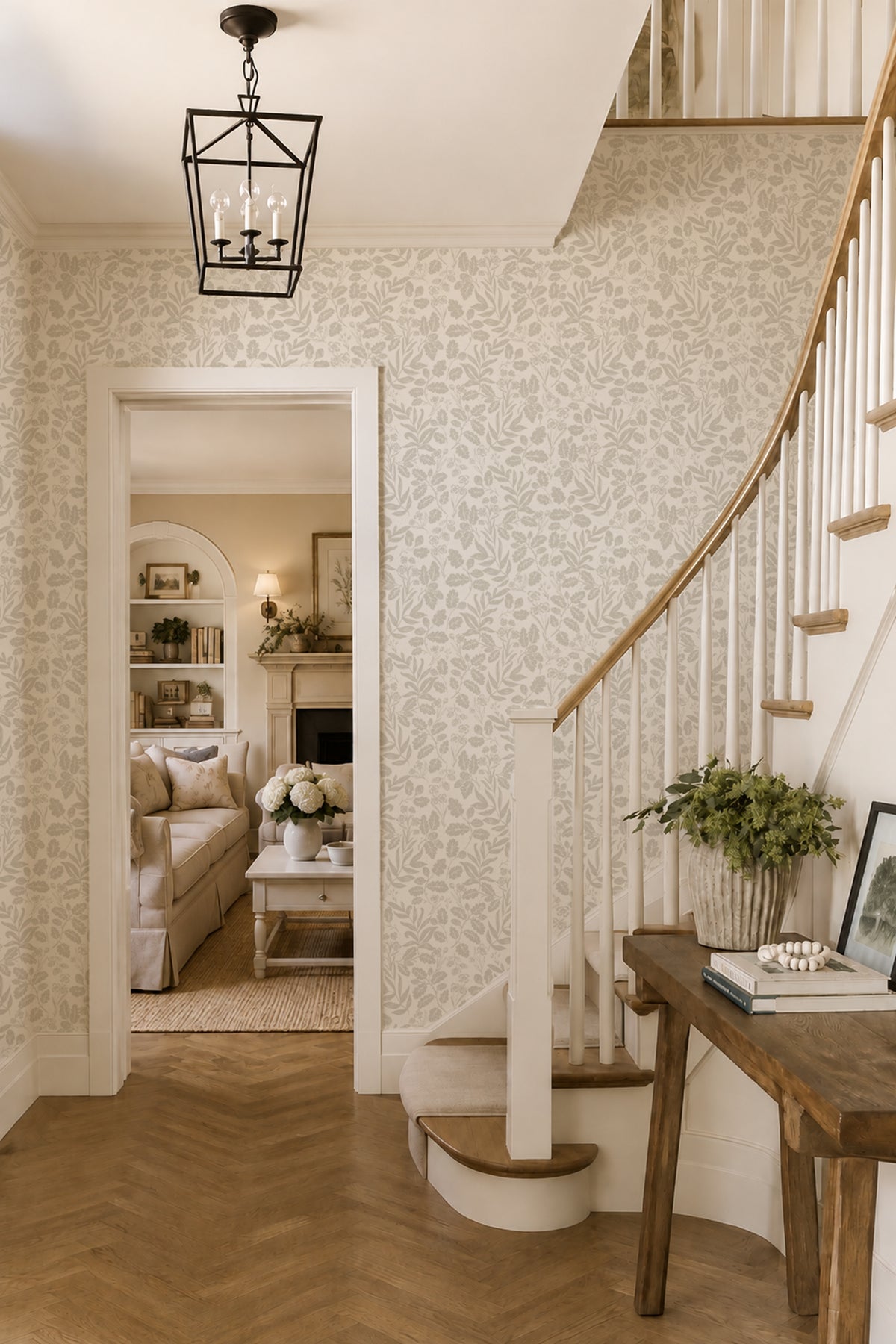 An elegant interior staircase with white wooden steps and a curved banister, set against a wall with light beige floral wallpaper. A dark wood console table sits at the base of the stairs, and a black metal lantern light hangs from the ceiling.