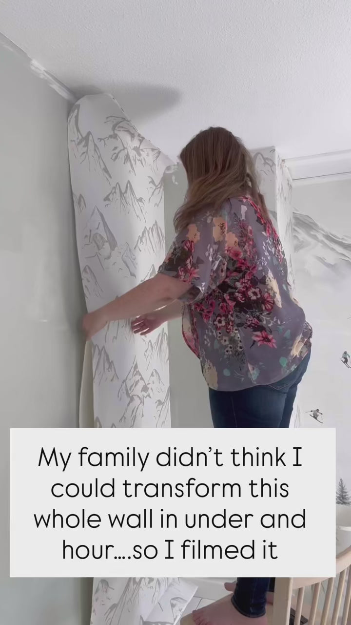 Load video: Neutral taupe mountain wallpaper on an angled wall during installation, with outlets exposed and white wood flooring.