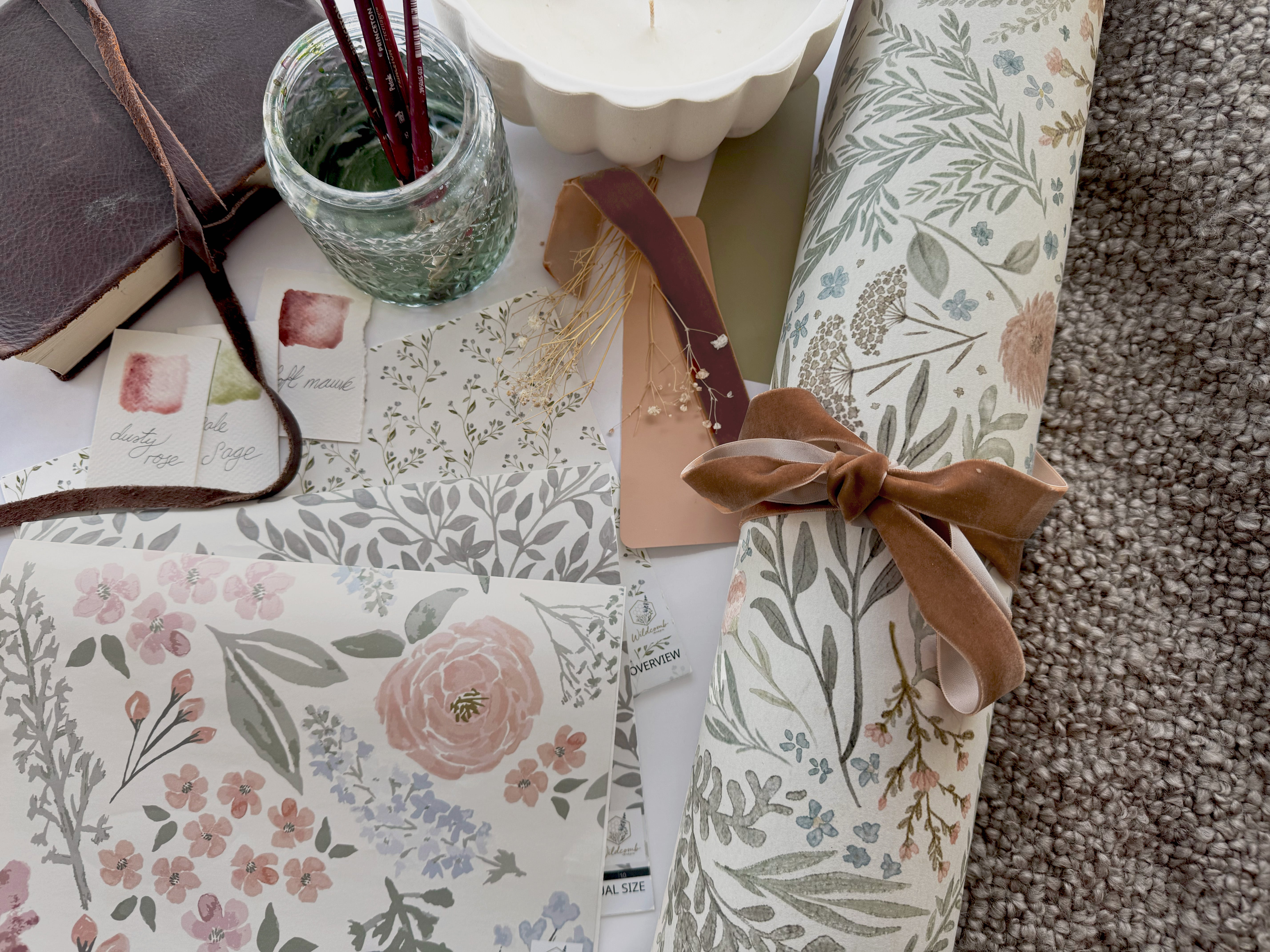 Rolled floral nursery wallpaper tied with a velvet ribbon, featuring hand-drawn blush roses, sage green leaves, and delicate wildflowers in soft watercolor tones — a romantic custom nursery décor design perfect for a baby girl’s room or personalized children’s wall art styling.
