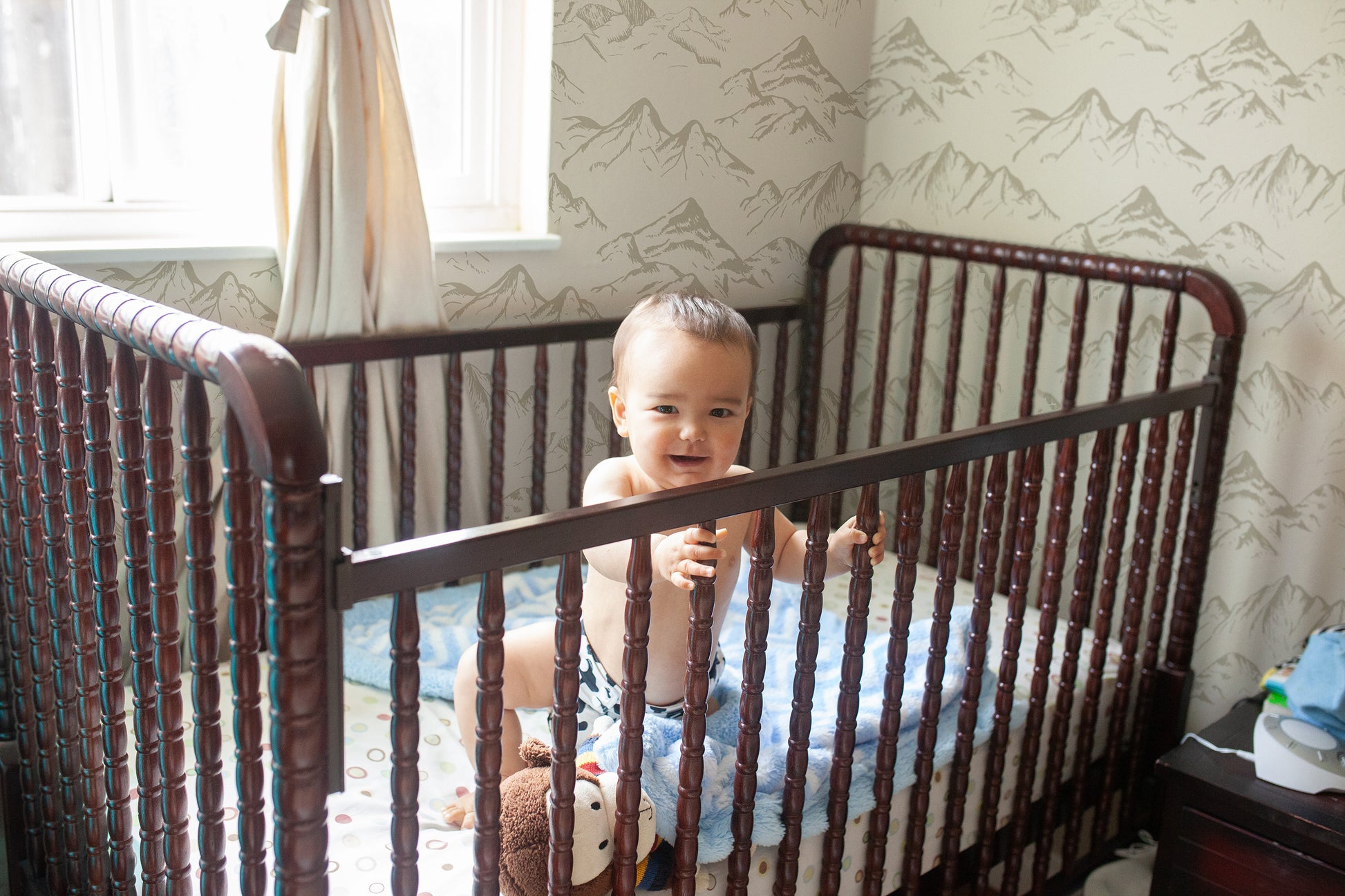 Baby standing in a dark wood crib with neutral taupe mountain wallpaper in a softly lit nursery.