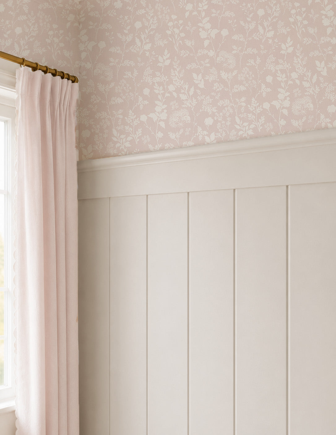 A styled interior corner showing pale blush pink floral wallpaper on the upper wall, white wainscoting below, a light pink curtain, and a gold curtain rod.