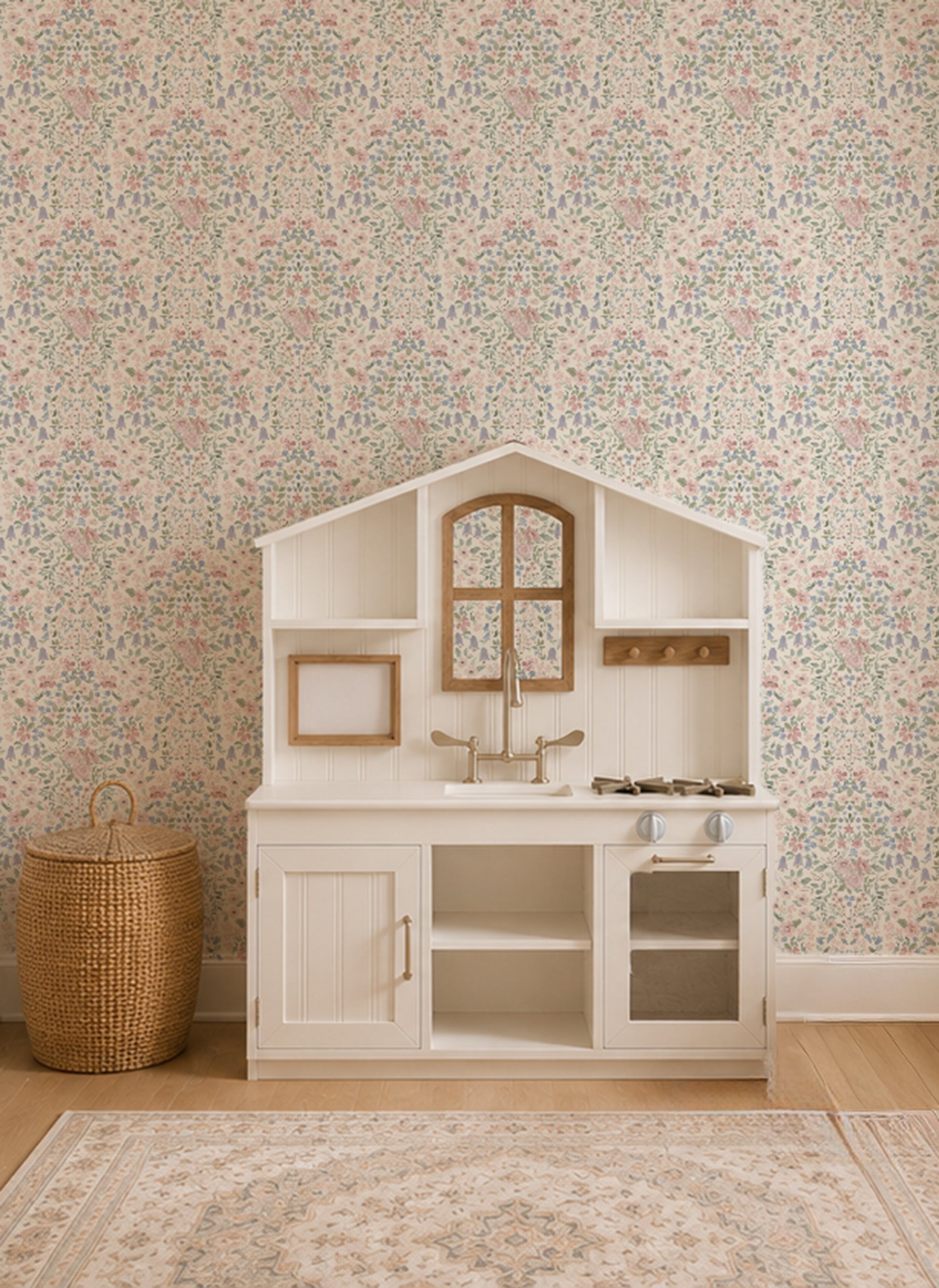A white children's play kitchen with a house-shaped hood, set against a floral wallpaper background. A woven basket sits on the wooden floor to the left.